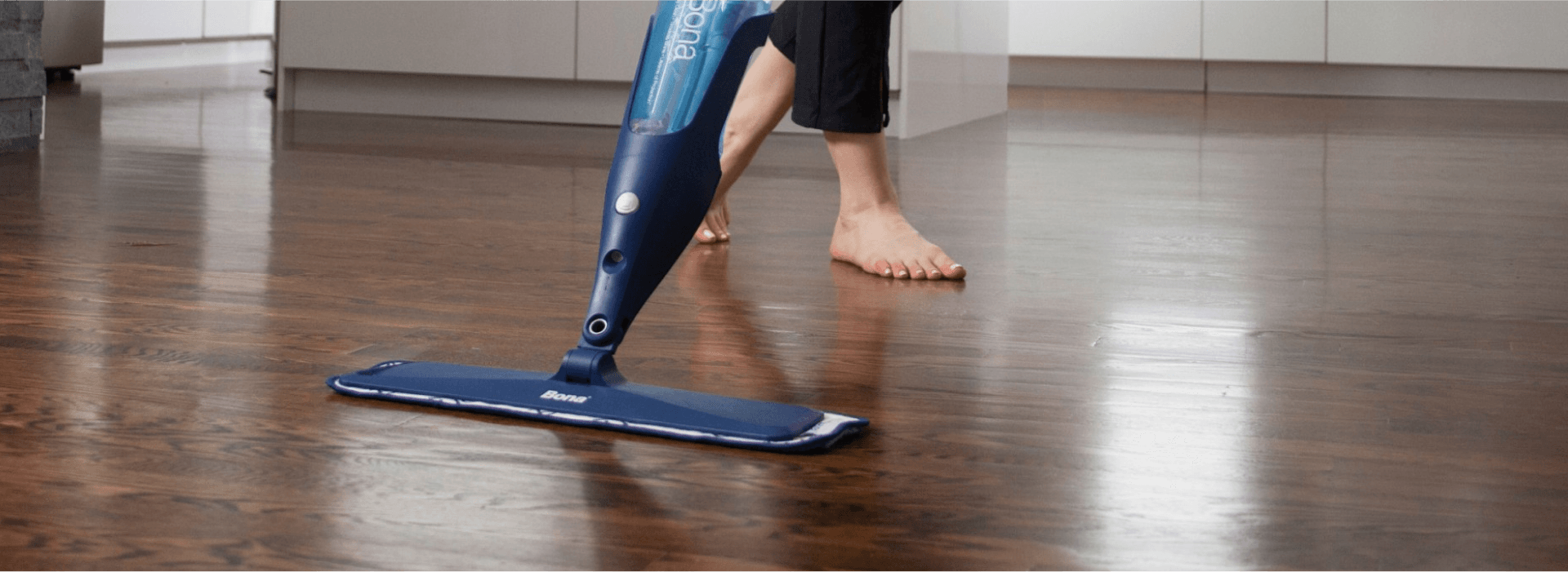 A barefoot woman in rolled up jeans cleans a hardwood floor in a kitchen with the Bona Spray Mop.