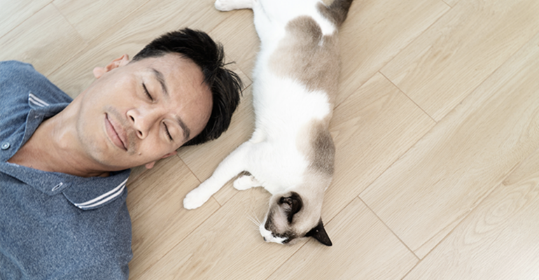 A smiling man lies peacefully on a clean hardwood floor with eyes closed. A gray and white cat lies next to his face.